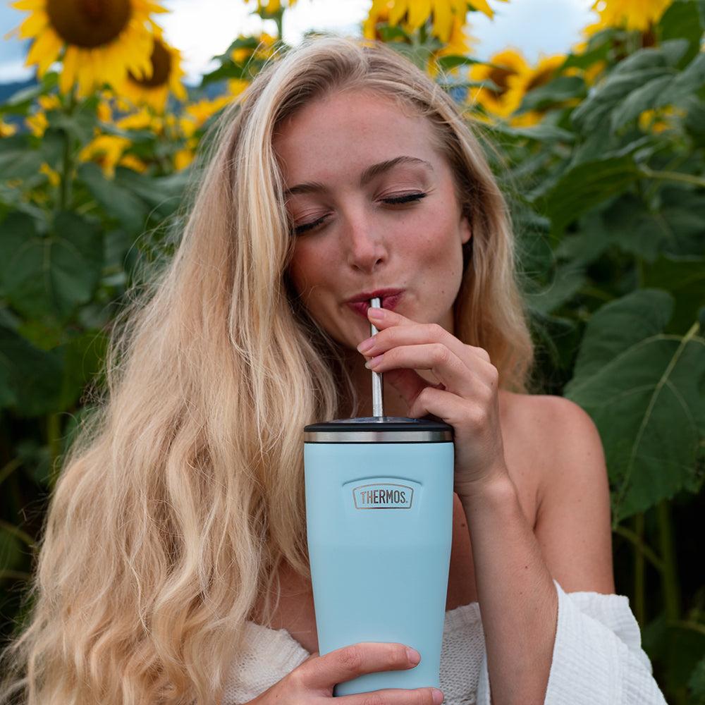 Woman standing in front of Sunflower field, drinking from a Thermos tumbler with straw.