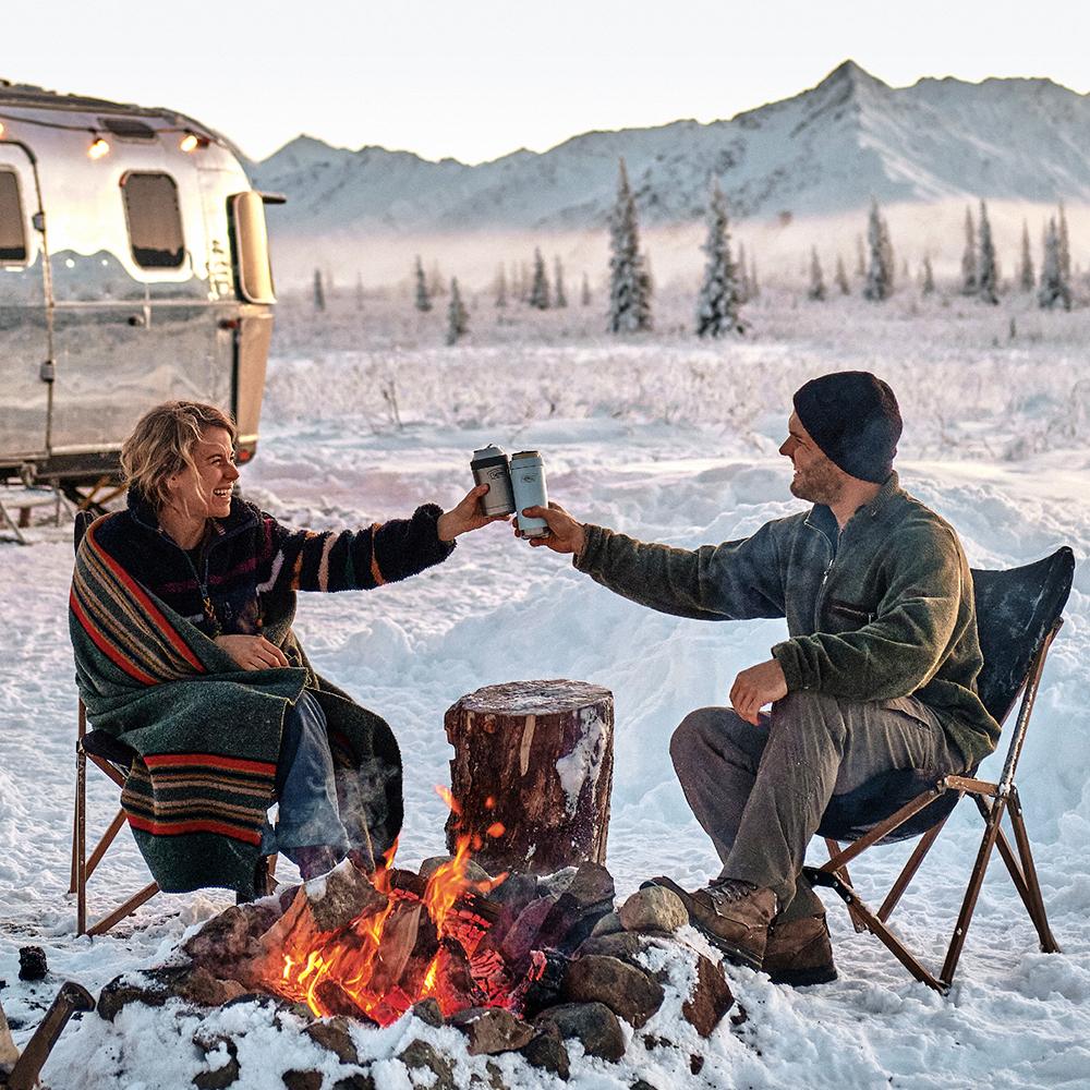 Man and woman in wintery outdoor scene, doing cheers with their Thermos Can Insulators.